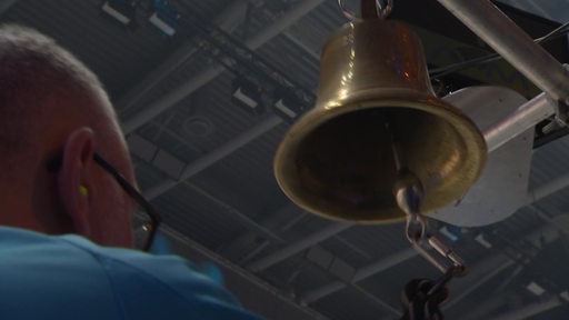 Ein Mann läutet eine große bronzefarbene Glocke in einer Halle. 