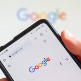 Eine Hand hält ein Smartphone, das den Google-Startbildschirm zeigt.