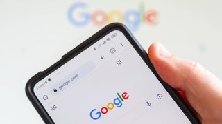Eine Hand hält ein Smartphone, das den Google-Startbildschirm zeigt.