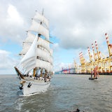 Ein großes und ein kleines Segelschiff fahren an Kränen eines Hafens entlang.