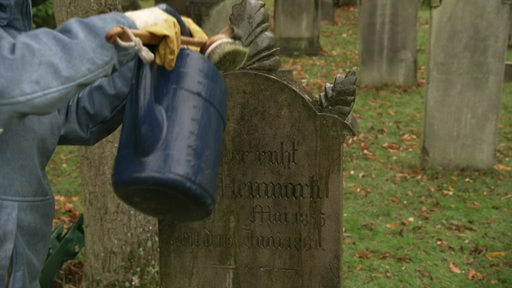 Jemand reinigt einen Grabstein auf einem Friedhof mit Gieskanne und einer Bürste.