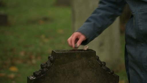 Eine Hand legt einen kleinen Stein auf einen Grabstein