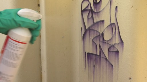 Eine Farbschmiererei an einer Wand wird mit Hilfe einer Spühflasche entfernt.