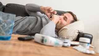 Ein Mann liegt krank auf einem Sofa und telefoniert mit einem Smartphone. 