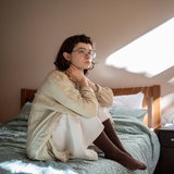 Eine junge Frau sitzt auf einem Bett und schaut grübelnd in die Luft.