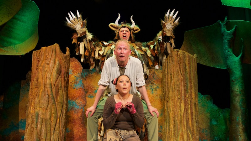Zwei Männer und eine Frau spielen in einer Waldlandschaft offenbar eine Theaterszene