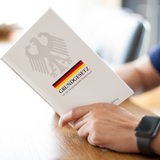Eine Person hält das Grundgesetz, GG, der Bundesrepublik Deutschland, in den Händen und liest darin an einem Tisch.