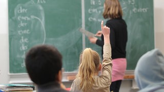 Die Lehrerin erklärt an der Tafel die Aufgaben, eine Schülerin zeigt auf