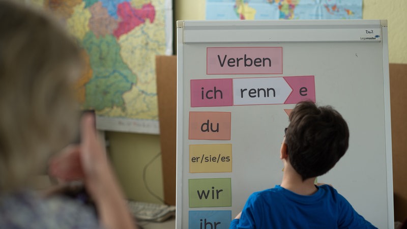 Ein Junge steht im Deutschunterricht vor einem Flipchart zum Thema Verben.