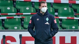 Werder-Trainer Florian Kohfeldt trägt eine FFP2-Maske und hält die Hände in die Jackentaschen gesteckt. 