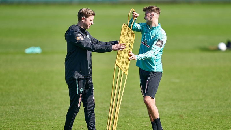 Werder-Trainer Florian Kohfeldt scherzt mit Niclas Füllkrug am Rande des Trainings und trägt mit ihm eine gelbes Sparrings-Metallmännchen.