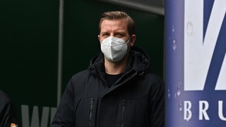 Florian Kohfeldt trägt Mund-und-Nasenschutz und steht im Weser-Stadion am Spielertunnel neben eriner großen Werbeflasche.