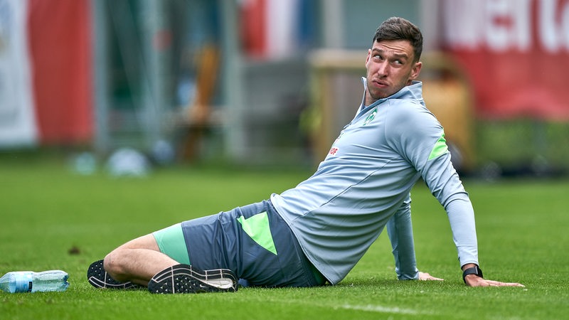 Werder-Torwart Jiri Pavlenka sitzt beim Training auf dem Rasen und dehnt sich etwas mit angespanntem Gesicht.