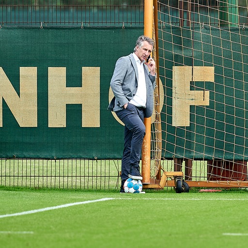 Werder-Sportchef Frank Baumann lehnt am Rande des Trainings an einem Tor und telefoniert.