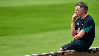 Werder-Sportchef Frank Baumann sitzt nachdenklich auf einer Bande und beobachtet das Training.