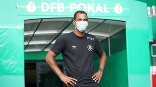 BSV-Trainer Benjamin Eta steht im Weserstadion am Augang des Spielertunnels mit freudigem Lächeln hinter der weißen Maske.