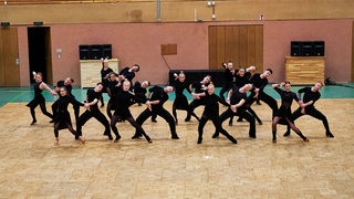 Die Lateinformation des Grün-Gold-Clubs bei der Präsentation ihrer neuen Choreografie in einer Sporthalle in schwarzer Trainingskleidung.