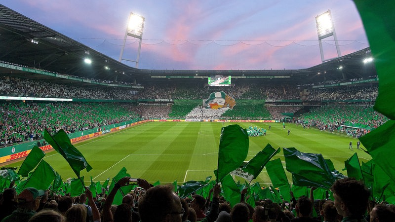 Blick in das vollbesetzte Weserstadion im Abendrot mit Fans, die grün-weißen Fahnen schwenken.