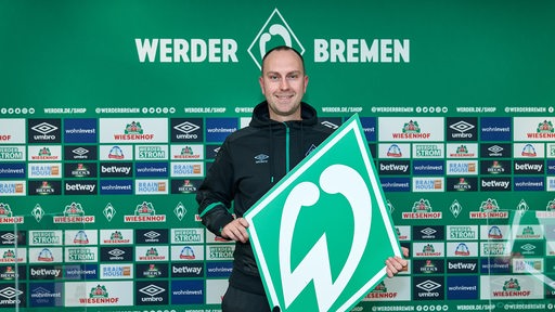 Werder-Trainer Ole Werner hält bei seiner Vorstellung strahlend eine große Papp-Werder-Raute in die Kamera.