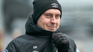 Werder-Trainer Ole Werner zieht sich auf dem Weg zum Trainingsplatz mit verschmitztem Lächeln den Jackenreißverschluss hoch.