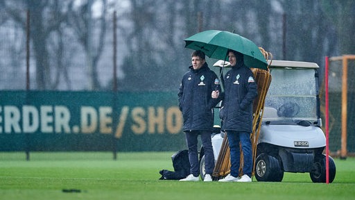 Frank Baumann und Clemens Fritz stehen am Rande des Trainings unter einem grünen Regenschirm im Nieselregen.