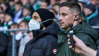Ömer Toprak mit weißer FFP2-Maske und Marco Friedl stehen vor der Ostkurve und verfolgen konzentriert das Werder-Spiel gegen Darmstadt.
