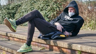 Marvin Ducksch lümmelt mit schwarzem Kapuzenpulli und Maske auf derSteintreppe und verfolgt das Werder-Training.