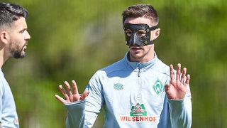 Werder-Profi Marco Friedl trägt eine schwarze Maske beim Training und streckt abwehrend die Hände nach vorne.