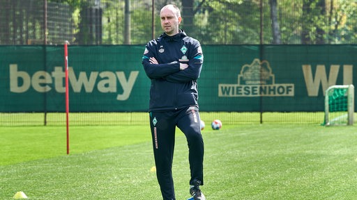 Werder-Trainer Ole Werner steht mit verschränkten Armen mit einem Fuß auf einem Ball und beobachtet das Training.