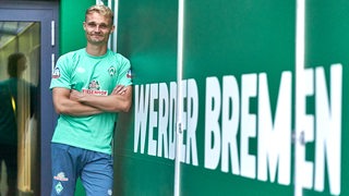 Amos Pieper posiert neben einem Schriftzug von Werder Bremen im Stadion.
