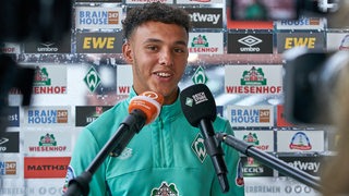 Werder-Neuzugang Lee Buchanan vor einer Werbewand mit Mikrofonen vor sich beim Interview.
