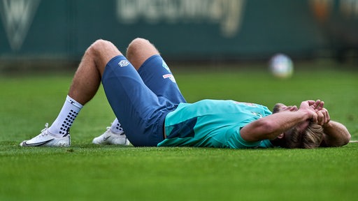 Werder-Stürmer Niclas Füllkrug liegt am Rande des Werder-Trainings platt auf dem Rücken auf dem Rasen und hält sich die Arme vors Gesicht.