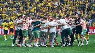 Werder-Spieler, Trainer und Betreuer liegen sich am Mittelkreis ausgelassen hüpfend in den Armen nach dem Sieg in Dortmund.