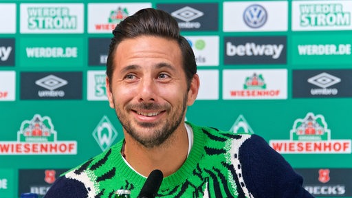Claudio Pizarro in einem auffällig-bunten Strickpullover grinst verschmitzt während einer Werder-Pressekonferenz.