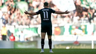 Werder-Stürmer Niclas Füllkrug von hinten mit ausgebreiteten Armen im Schatten auf dem Spielfeld in Freiburg.