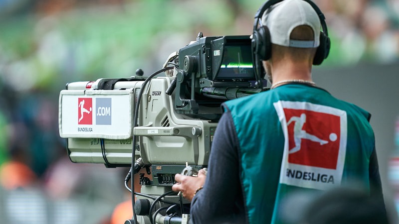 Ein Mann mit dem Logo der Bundesliga auf der Jacke sitzt am Spielfeldrand an einer TV-Kamera.