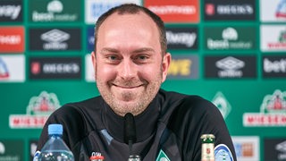 Werder-Trainer Ole Werner mit breitem Lächeln bei einer Pressekonferenz.