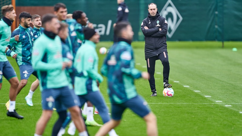 Werder-Coach Ole Werner beobachtet konzentriert und mit verschränkten Armen das Aufwärmen seiner Spieler beim Training.