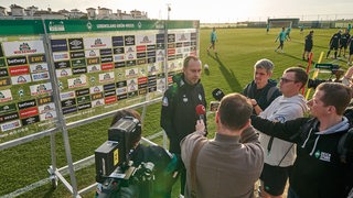 Werder-Trainer Ole Werner am Rande des Trainingsplatzes im spanischen Murcia vor einer Werbewand beim Interview.