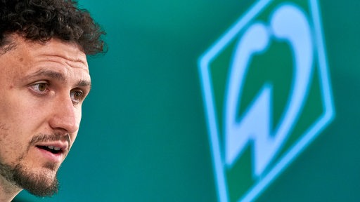 Werder-Verteidiger Milos Veljkovic in Nahaufnahme von der Seite bei einer Pressekonferenz, hinter ihm an der grünen Rückwand eine Werder-Raute.