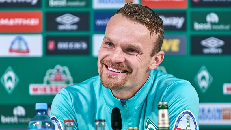 Werder-Spieler Christian Groß strahlt auf dem Podium einer Pressekonferenz.