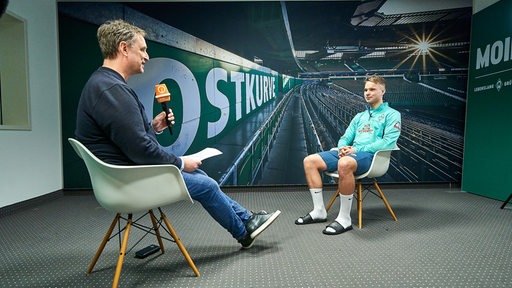 Sportblitz-Reporter Stephan Schiffner sitzt auf einem Stuhl dem Werder-Spieler Niklas Schmidt beim Interview gegenüber, hinter ihnen eine Fototapete der Ostkurve im Weser-Stadion.