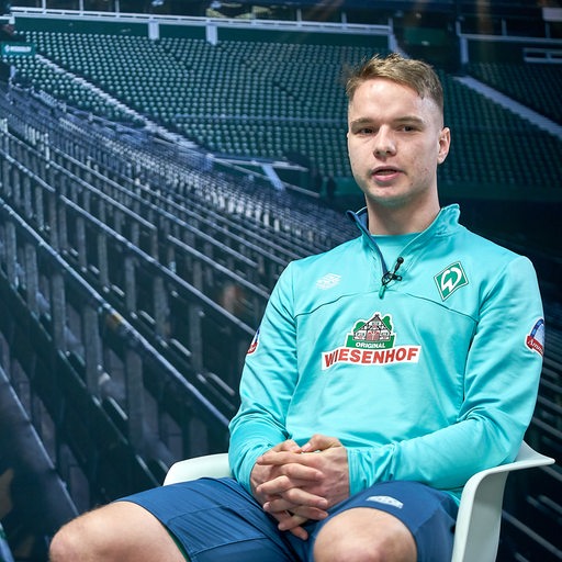 Werder-Profi Niklas Schmidt sitzt bei einem Interview vor einer Fototapete, die einen Blick ins Weser-Stadion zeigt.