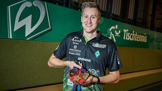 Werders Tischtennis-Spieler Mattias Falck posiert strahlend mit seinen WM- und EM-Medaillen vor einer grünen Bande mit Werder-Logo und dem Schriftzug "Tischtennis".