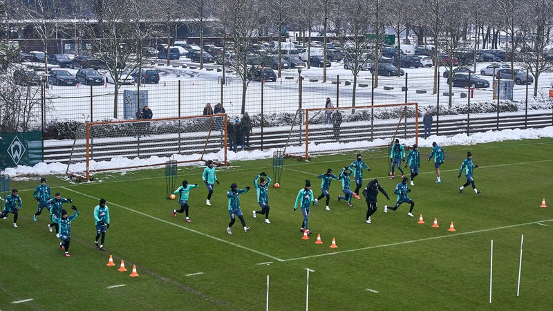 Blick von oben auf den Trainingsplatz vor dem verschneiten Weser-Stadion, Werder-Spieler wärmen sich auf dem freigeräumten Rasen auf.