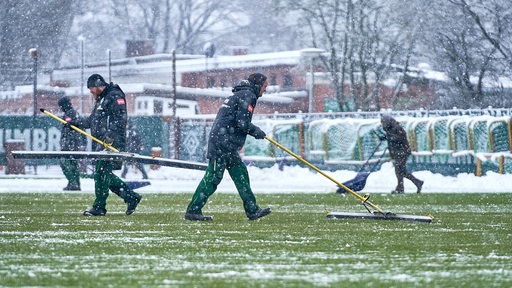 Mehrere Helfer schippen im Schneetreiben auf dem Trainingsplatz vor dem Weser-Stadion den Schnee weg.