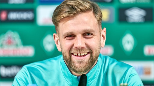 Werder-Stürmer Niclas Füllkrug mit breitem Grinsen bei einer Pressekonferenz, seine markante Zahnlücke blitzt dabei auf.