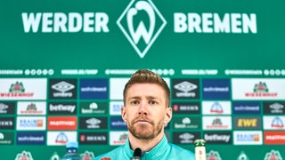 Werder-Spieler Mitchell Weiser sitzt regungslos auf dem Podium einer Pressekonferenz vor einer Werbewand.