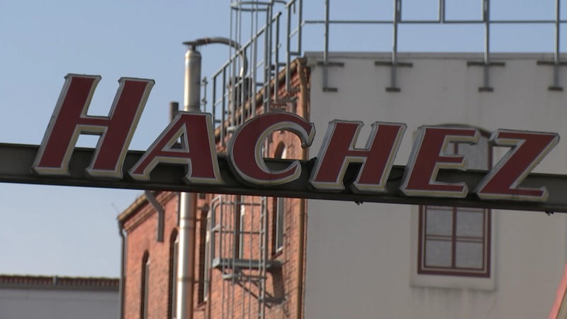Ein Schild mit dem "Hachez"-Logo, im Hintergrund ist das Gebäude der Schokoladenfabrik zu sehen.