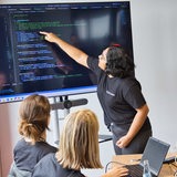 Eine junge Frau erklärt auf einem großen Screen anderen Frauen einen Computer Code